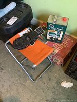 Foldable orange camping stool with metal frame, Coleman camp fuel can, and Life Sports black ice cleats placed on and around the stool.
