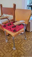 Two vintage kitchen chairs shown from an angle, one with tan vinyl upholstery and removable red floral cushion, the other wooden with simple backrest.
