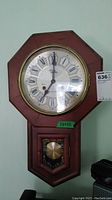 Clear front view of wooden octagonal Style House wall clock with visible clock face, Roman numerals, and brass pendulum behind glass panel.