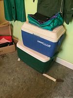 Two stacked hard coolers: top blue and white Coleman Polylite 40 cooler, bottom dark green and white Igloo cooler, teal cooler bag on top.