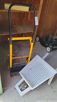 Utility dolly with black and yellow frame standing upright, wheelbarrow lying on the ground.