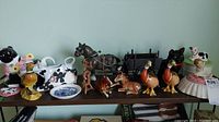 Wide view of shelf showing various ceramic figurines including cow teapot, cream pourer, duck trinket boxes, and horse and carriage figurine.