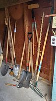 Photo showing rake, several shovels with wooden handles, a pick, an ax, and a large wooden auger hanging on a wall inside a shed