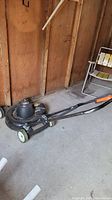 Side view of black Black and Decker electric lawn mower with handle and wheels in a wooden shed.