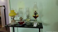 Four lamps on black table against wall, including three oil lamps and one yellow glass electric lamp with cord.