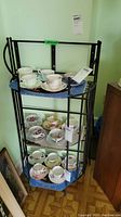 Black metal corner shelf unit displaying china cups and saucers, blue shelves, positioned in room corner.