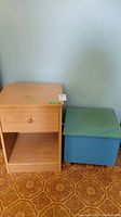 Wooden bedside table and blue chest side by side on a patterned floor against a plain wall.