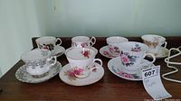 Full view showing all seven tea cups and saucers arranged in two rows, illustrating variety of floral patterns and gold accents.