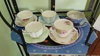 Photo showing five assorted tea cups with matching saucers arranged on a blue paper with decorative edges on a small shelf.