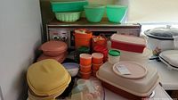 Overview of a variety of vintage Tupperware containers in multiple colors including green, orange, yellow, pink, red, and white stacked on a stove and nearby surfaces.