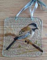 Clear textured glass suncatcher with a painted bird (chickadee) perched on a branch. Attached light blue ribbon for hanging.