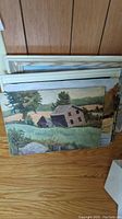 Stack of 16x12 landscape paintings on wood floor with two white wooden frames behind