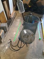 Photo of large galvanized metal trash can with lid, showing some rust, with items inside including bird seed bags and pole. Two large black steel hooks positioned in front.