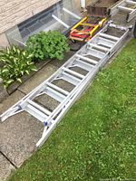 Overall view of the metal multi-purpose ladder laid on pavement next to grass and plants, with the hand cart placed above it, showing the condition and detailed structure of both items.