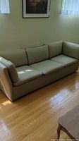 Beige three seater couch with high arm rests and short back, placed on wooden floor next to window with light coming in.