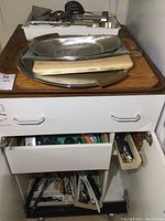 Photo of cabinet with cutlery trays inside drawers, assorted kitchen utensils, electric carver parts, and wooden cutting boards with metal trays on top.