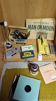 Overview of vintage household items including newspaper, telephone, iron, tins, coffee grinder, records, ice tray, and lamp displayed on wooden surface