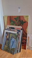 Stack of original unframed paintings leaning against a wall showing plant still life and river scene artworks visible