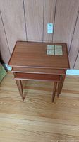 Full view of the three nesting teak side tables stacked together against a wood paneled wall with tile inset on the largest.