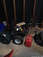 Front-left view of green Craftsman 27-inch gas snowblower parked indoors showing auger housing, wheels, and handles with red fuel can at right and operator manual on belt cover.