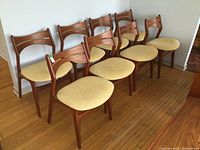 Eight teak chairs arranged in a row against a wall on hardwood floor