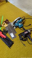 Photo of the entire lot on yellow carpet, includes Makita circular saw, Craftsman electric drill, two hand saws, and Black & Decker shake light with charger and black bag.