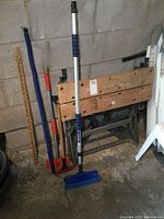 Wooden folding workmate stand with two car wash brushes and a wooden yardstick leaning against a wall in a basement or workshop area.