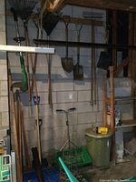 Various garden and yard tools hanging on wall and stored against it including shovels, rakes, trays, electric weed eater and tomato cages