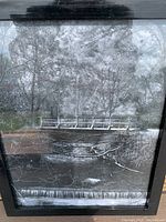 Black and white framed picture showing a winter or frosted nature scene with a bridge over a river or creek and frost-covered trees
