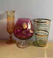 Photo shows three glass vases: amber ruffled rim vase, cranberry vase with gold floral details, and green glass vase with gold stripes.