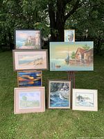Group view of all 8 paintings displayed outdoors leaning against a fence and tree, showing various landscape styles in different sized frames