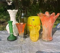 View of all seven glass vases displayed together including white milk glass vase, yellow and orange Marc Aurel vase, green frosted glass piece, and orange-clear vase.