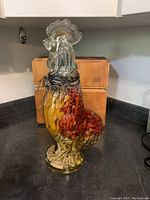 Front view of clear glass rooster bottle filled with yellow, red, and white pickled peppers, sits on a black countertop with wooden boxes behind