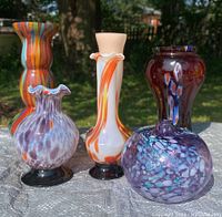 Six spatter glass vases grouped on textured surface with outdoor background. Various colors including orange, blue, purple, and red. Shows shapes and sizes of vases.