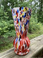 Front view of multicolored spatter glass vase on wooden railing with foliage background.