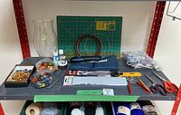 View of one shelf showing multiple craft items including a rotary mat, Mastercraft cordless cutter in blue case, beads, paintbrushes, glue gun, and cutting tools.