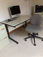 Desk and chair setup with an iMac computer and mouse shown on the desk. Desk has a light colored surface, black metal frame and adjustable legs. Chair is gray fabric on caster wheels.