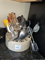 Photo of assorted cooking utensils including wooden, metal, and plastic ones stored upright in a cream utensil holder on countertop.
