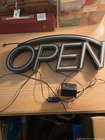 The electronic OPEN sign shown from the front, displayed on a wooden surface along with the power cord attached.