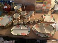 Full view of the dining set on a table showing the various plates, divided dishes, bowls, cups, and teacups all featuring the pink flower and green leaf design.