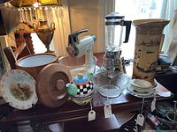 Full view of the assorted kitchenware items on wooden table including blender, bowls, plates, teapot, and decorative pitcher.