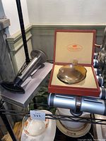 Kitchen items on wire rack including stainless steel pineapple corer, small decorative dish in box, and paring cutter.
