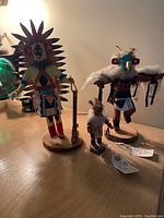 Four Kachina dolls of different sizes displayed on a wooden surface, showing hand-painted details and feather adornments.