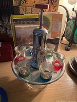 Shot distributor with 6 shot glasses on round metal tray, plunger and spout visible