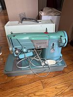 Full front and side view of vintage Singer sewing machine with foot pedal and power cord attached, sitting on wooden floor with white hard case behind.
