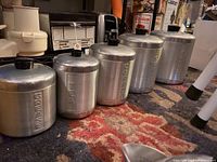 Five labeled stainless steel storage containers on a carpet, labeled Grease, Tea, Coffee, Sugar, with an additional unlabeled container.