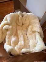 Photo showing the exterior of the cream faux fur coat laid open in a box on a wooden floor.
