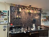 Wide view of bar area with shelves holding beer mugs, stemware, steins, and a row of vintage beer cans lined up on a shelf mounting on black tiled backsplash.