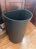 Black triangular plastic trash bin with smooth matte finish, shown standing on wood flooring near cabinetry.
