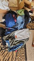 Photo showing blue duffle bag with assorted folded pants including olive green pants and jeans on a patterned carpet with some boxes in the background.